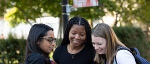 Three students smiling and looking at a smartphone outside.