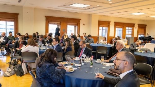 People attending the conference are seating in round tables. Professor Perrone is seen seating down and holding a glass.