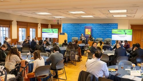 People attending the conference seating at round tables. In the background, there is the stage with the speakers.