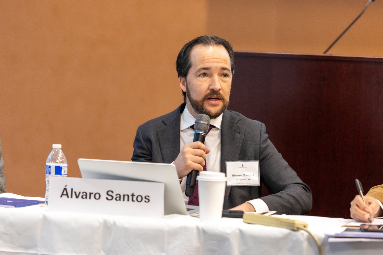 Professor Alvaro Santos holding a microphone while speaking seating at a table