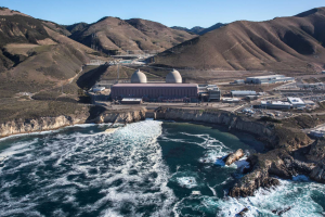 A bird’s eye view of the Diablo Canyon Nuclear Power Plant with the blue waves of the Pacific Ocean seen crashing on rocks in the foreground, brown rolling hills and a blue sky are seen in the background.