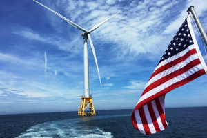 A white offshore wind turbine with a yellow platform is seen in the center of the image rising out of the deep blue ocean. The wake of a boat, the American flag flying on a boat, and a blue sky with fluffy clouds are also visible.