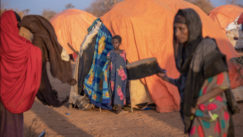 Child stands in front of an orange tent in the desert while robed women walk by.