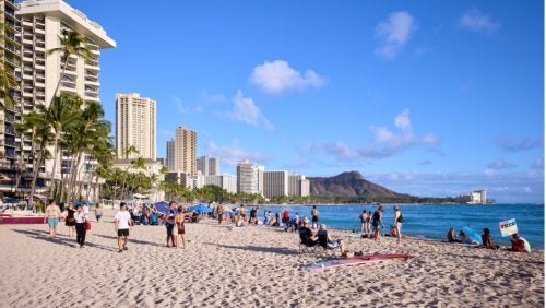 Waikīkī Beach on a sunny day, with a number of visitors on the sand and near the water.