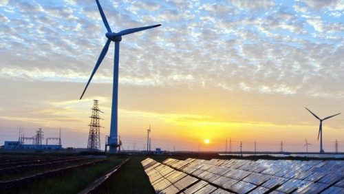 Solar cell panels in the foreground, wind turbines in the middle ground, and electricity pylons in the background at sunset
