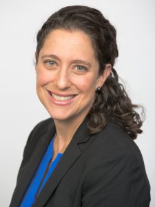 A woman with dark, curly hair and a blazer smiles at the camera. 