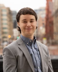 Man with brown hair wearing grey suit with blue shirt in front of city setting.