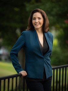 Woman with brunette hair and teal blazer leaning on black fence outdoors