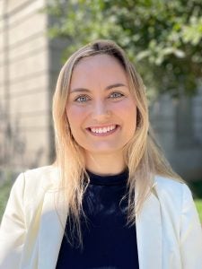 Melody Vidmar in white blazer in front of green trees.