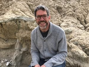 Image of David McNeill smiling and wearing a grey sweater while sitting on tan rocks.