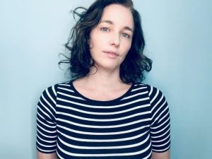 Image of Emily Tucker wearing a horizontally striped shirt and standing against a light blue wall.