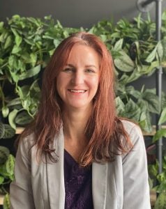 Headshot of a woman with red hair in front of a green foliage background