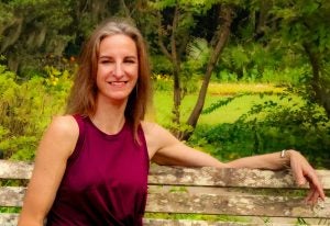 Smiling Woman with long brown and grey hair in sleeveless burgundy dress seated on an old brown bench, surrounded by trees and lush green foliage