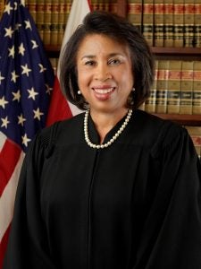 A smiling woman in a judges robe in front of the American and D.C. flags.