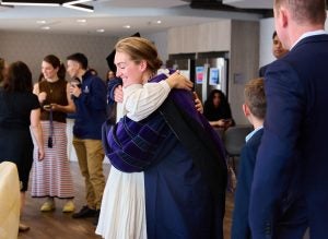 A graduate, wearing their regalia, hugs another individual