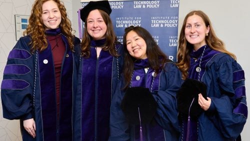 4 graduates, wearing regalia, smile in front of a Institute of Technology Law and Policy sign.