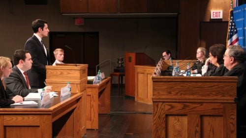 Side view of a student speaks at a podium in front of a panel of judges, co-counsel, and opposing counsel. 