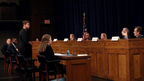 Student speaks at a podium in front of a panel of judges, co-counsel, and opposing counsel. 