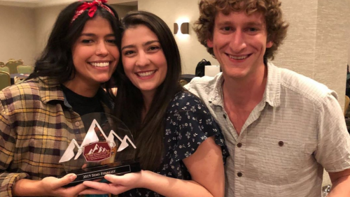 Three students, casually dressed, smiling holding awards. The awards are shaped like mountains and are clear.