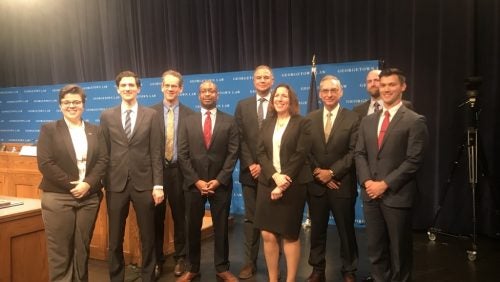 Nine people gathered together smiling. Edge of the photo has a courtroom set up and a blue banner with the words georgetown law written across it.