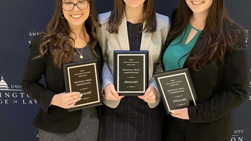 Three students, all wearing glasses, holding three plaques that read the word finalist on it. They are standing in front of a blue background with the words American University Washington College of Law