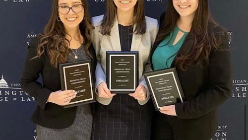 Three women smiling with plaques in front of American University Washington College of Law Banner