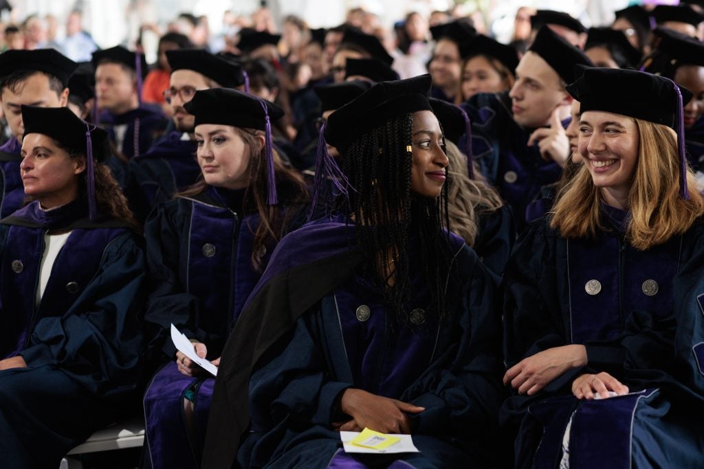 Two graduating students smile at each other, sitting with graduating classmates