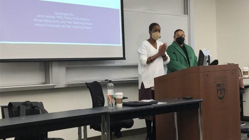 Professor Janel George and Clinical Teaching Fellow Antonio Coronado standing at a podium with a presentation slide on the screen behind them. 