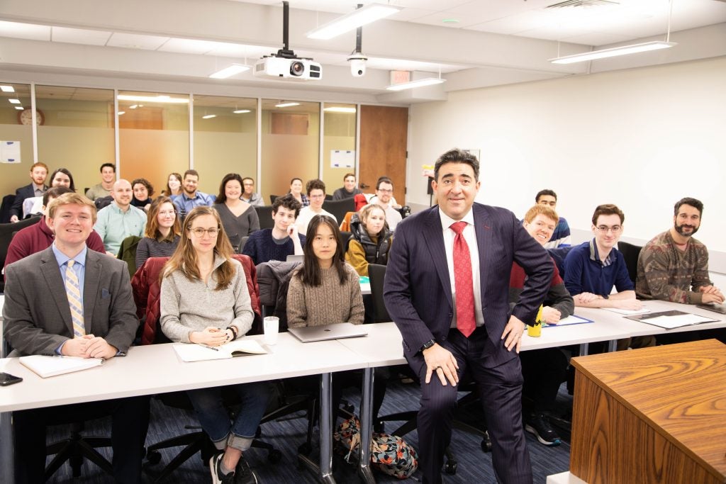 A professor wearing a suit and red tie in the classroom with four rows of students seated behind him.