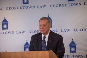 Senator Dick Durbin, wearing a dark suit and blue necktie, speaking at a podium