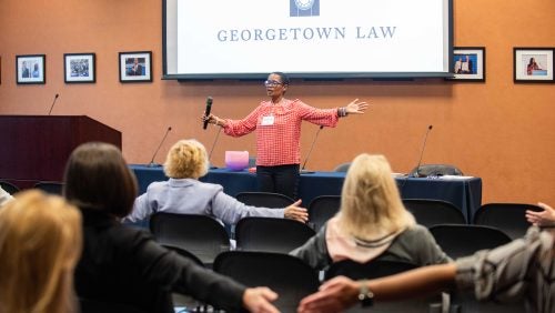 Ananda Leeke L' 91 leads a mindfulness workshop for a group of women with her hands raised in front of the room.