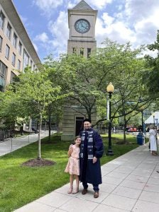 A graduating student in regalia standing on the Georgetown Law campus with a young girl