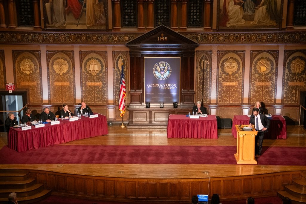The stage at Georgetown University's Gaston Hall, set up for a Faculty Moot Court demonstration, with a man standing at the podium to introduce the other participants seated at tables