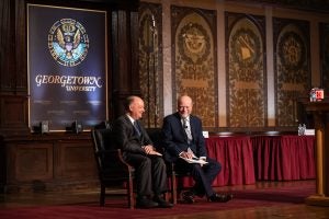 Two men, wearing suits, sitting together on stage in Georgetown University's Gaston Hall