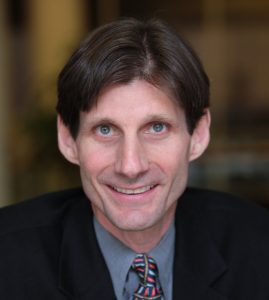 Image of a white man with dark hear, wearing a jacket and tie