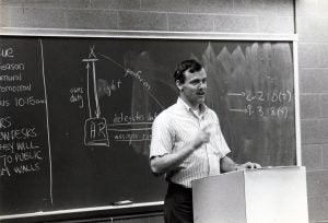 A man standing at a classroom lectern, with a blackboard on the wall behind him
