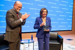Dean William M. Treanor, wearing a suit and applauding, standing next to Rep. Nancy Pelosi, who is holding a glass trophy.