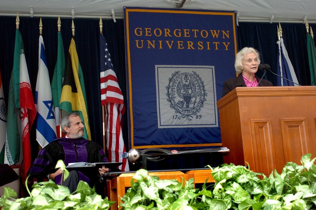 A man in academic regalia, seated on stage, looking at Justice Sandra Day O'Connor, who is speaking at a podium