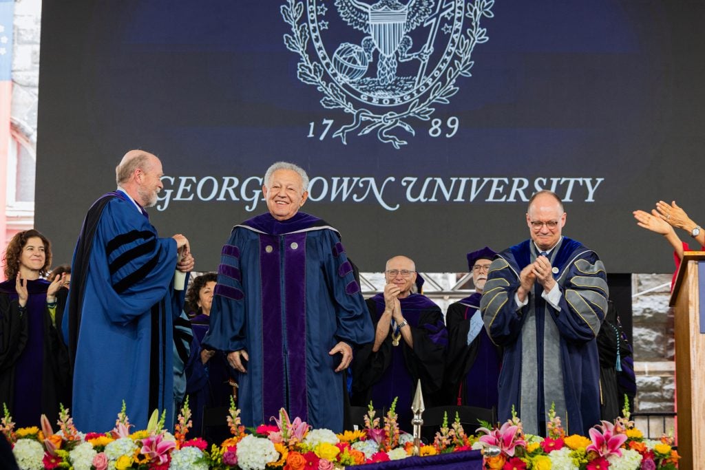 An image of the stage at the 2024 Georgetown Law commencement celebration, as faculty and administrators applaud for a man standing in academic robes.