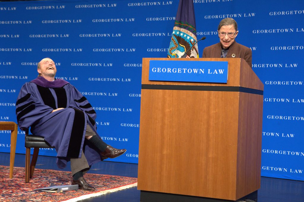 A man sitting in a chair, laughing, next to Supreme Court Justice Ruth Bader Ginsburg standing at a podium