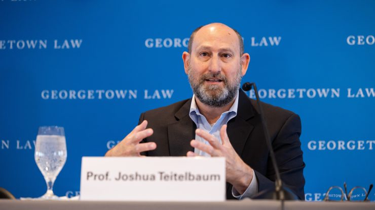 A bearded man sitting at a table speaking into a microphone. A printed card identifies him as Prof. Joshua Teitelbaum.