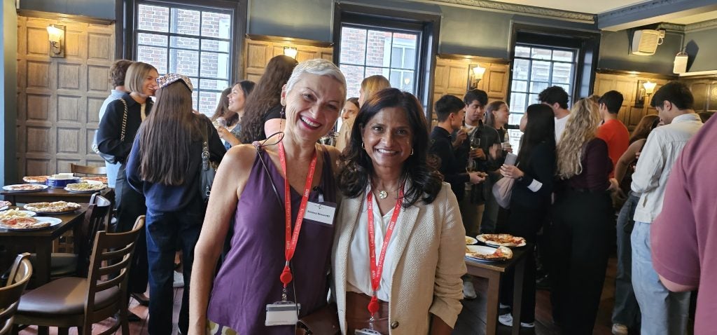 Two women smiling side by side, in a pub with a group of younger people in the background