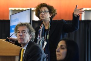 A woman wearing glasses, standing and speaking at an event, with two people seated in front of her