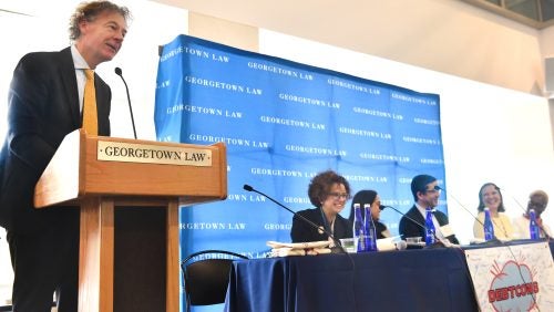 A man speaking at a podium, with a panel of people seated at a table beside him