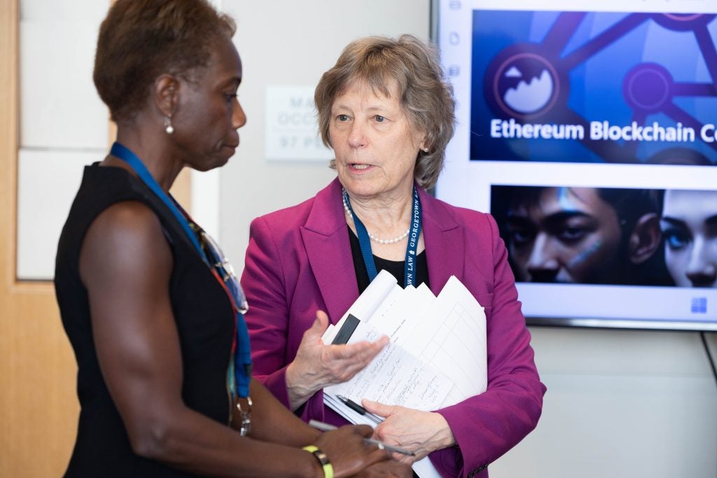 Two women stand together in a meeting room and converse with one another.