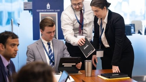 Three students, two standing and one sitting, cluster together in a meeting room during the Sim