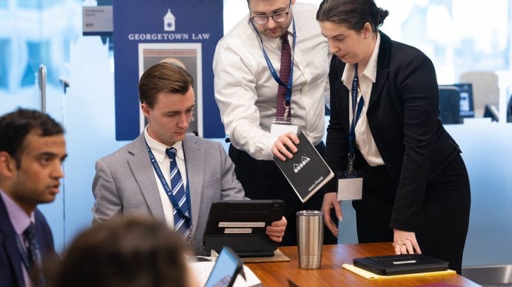 Three students, two standing and one sitting, cluster together in a meeting room during the Sim
