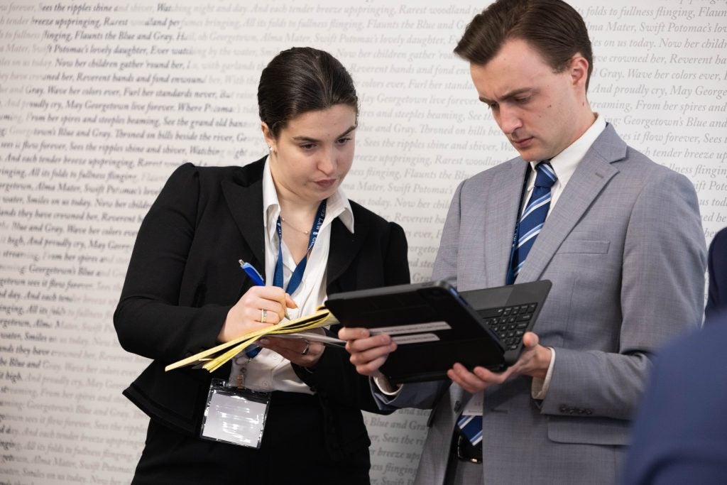 Two students stand together, focusing intently. Both look at the tablet screen one is holding while the other takes notes on a legal pad.