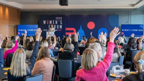 A room full of attendees pictured sitting at tables from behind, raising their hands.