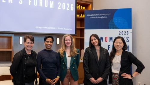 Five women stand and smile in front of a Women's Forum sign in a meeting room.